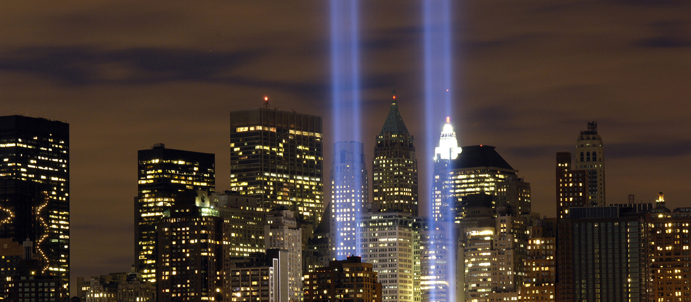 Two vertical beams of light shine upward among tall buildings in a city at night.