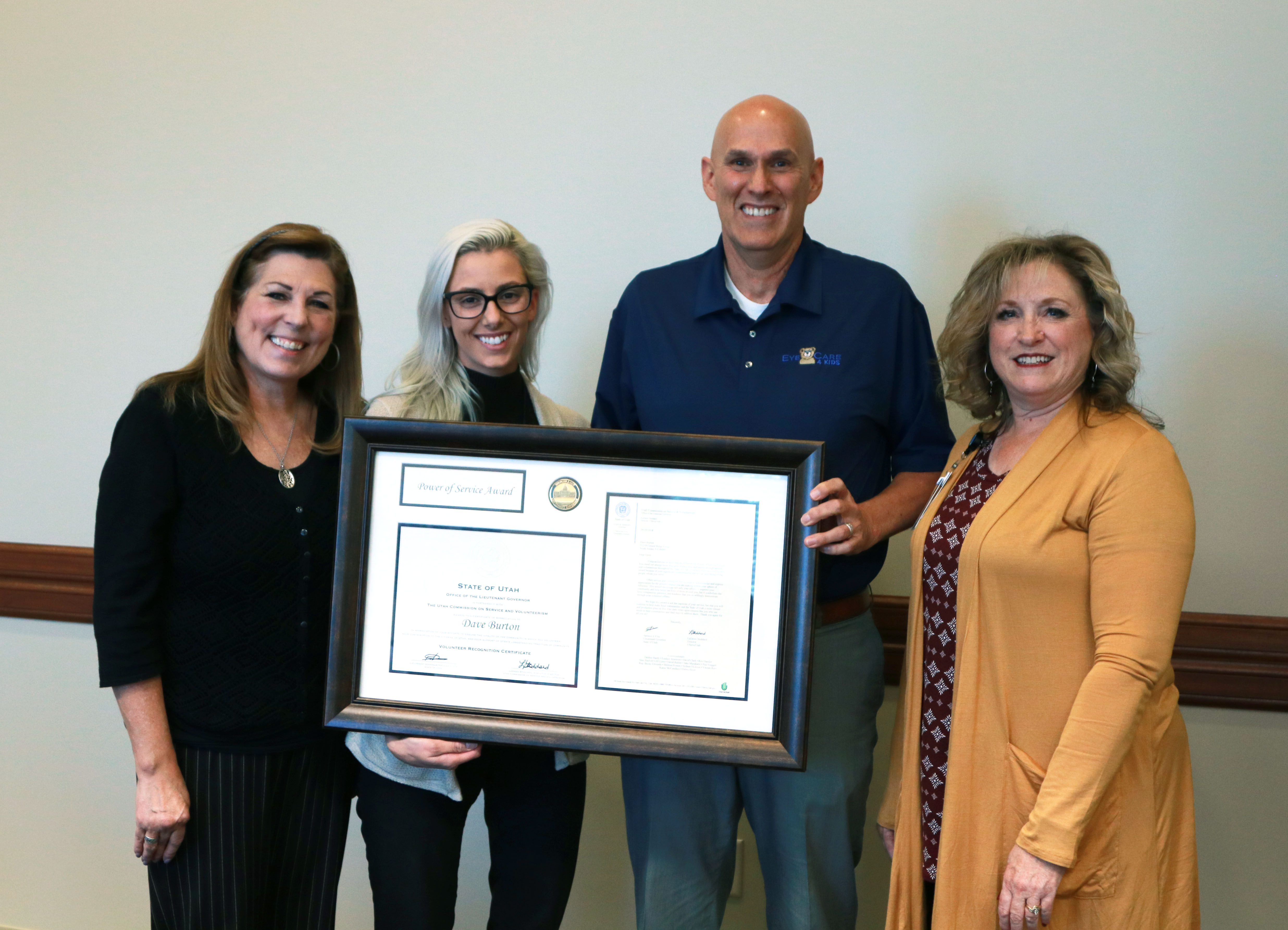 Four people standing indoors, holding a framed certificate and smiling at the camera.