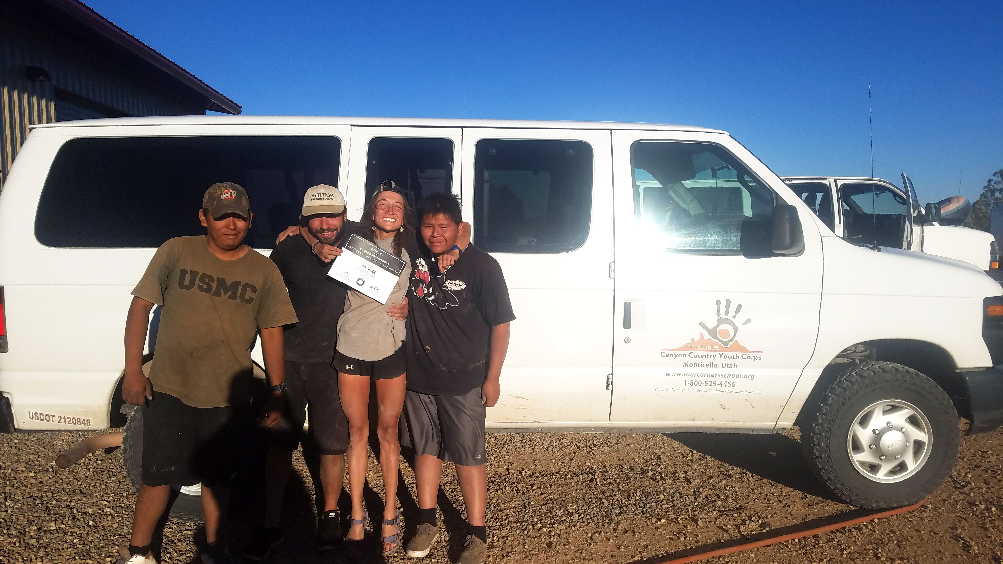 Four people stand smiling in front of a white van, one holding a certificate.