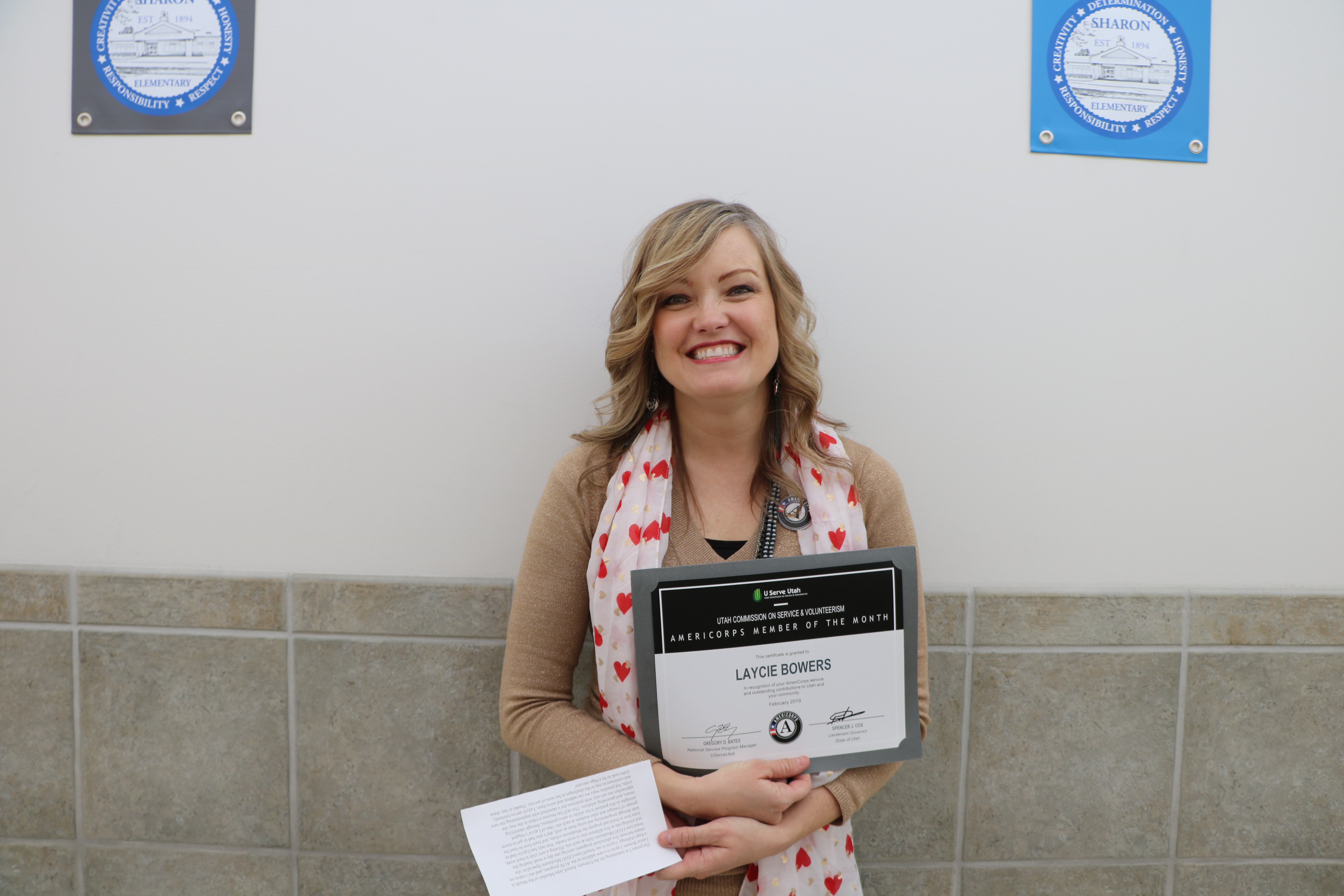 Woman smiling while holding a certificate and a piece of paper against a tiled wall.