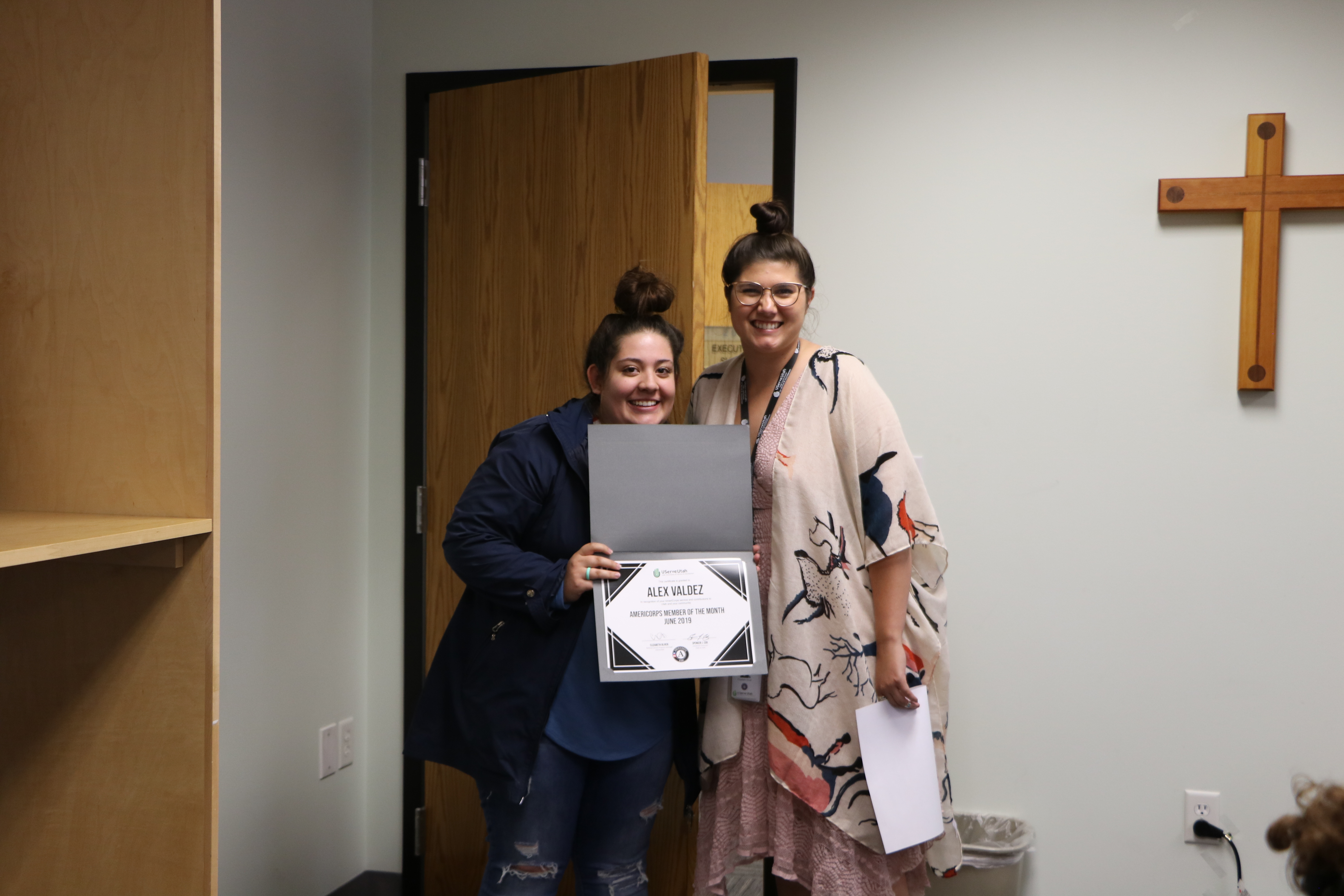 Two women stand together; one holds a certificate while the other holds papers and smiles.