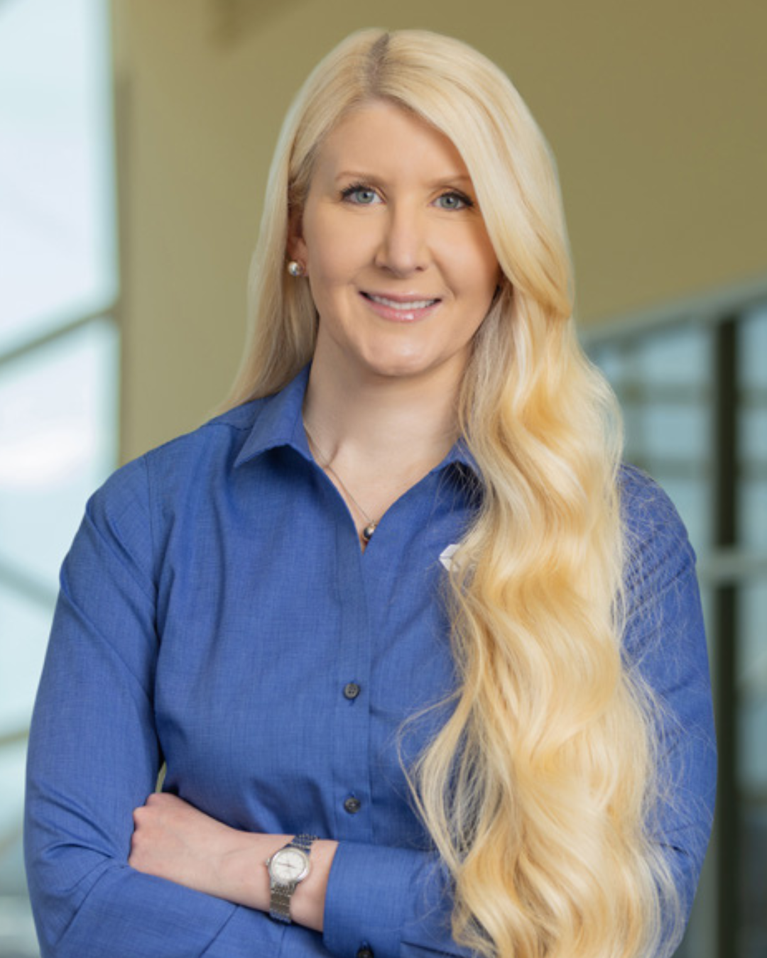 Woman with long wavy blonde hair smiles at camera with arms folded wearing blue collared shirt in indoor space