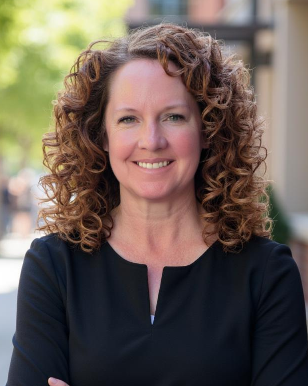 Woman with shoulder length curly brown hair smiles at camera wearing black top in outdoor setting