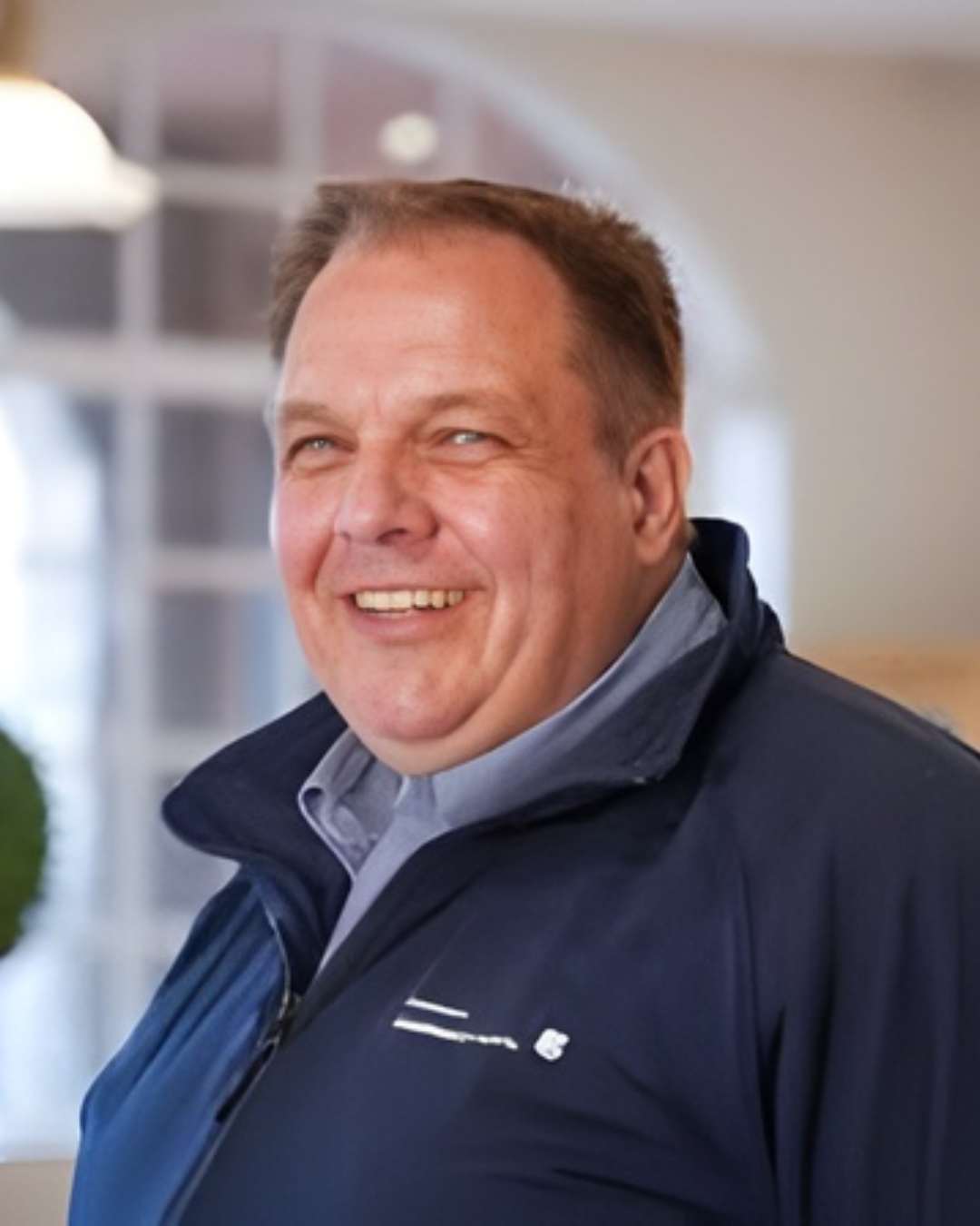 Man with short hair smiles at camera wearing navy jacket in indoor setting