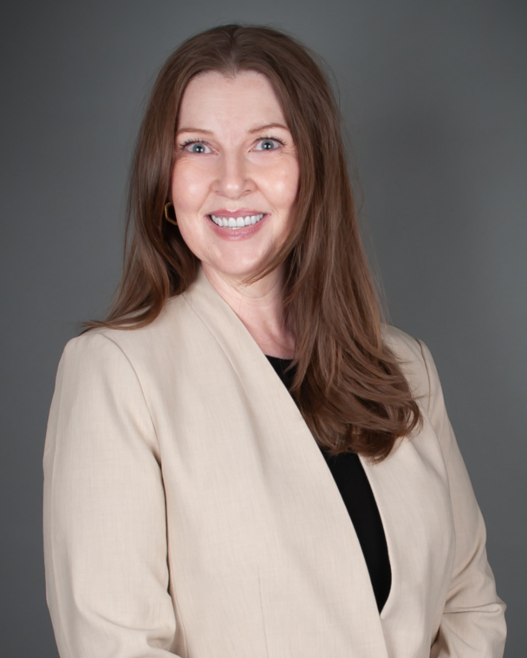 Woman with long brown hair wearing light tan blazer and dark top smiles at camera in front of gray background