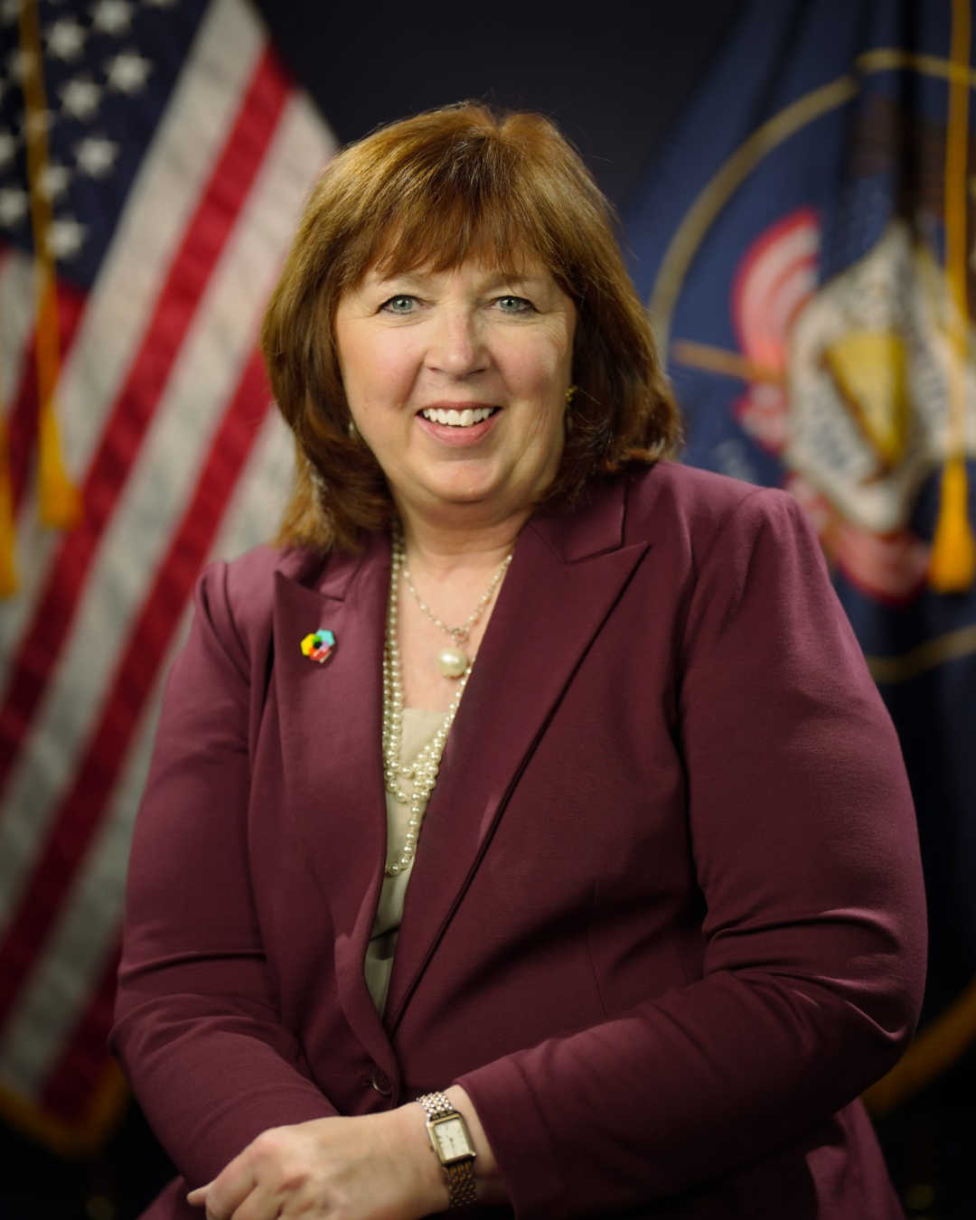 A woman in a maroon suit sits and smiles in front of the U.S. and Utah state flags, with her hands resting in her lap.