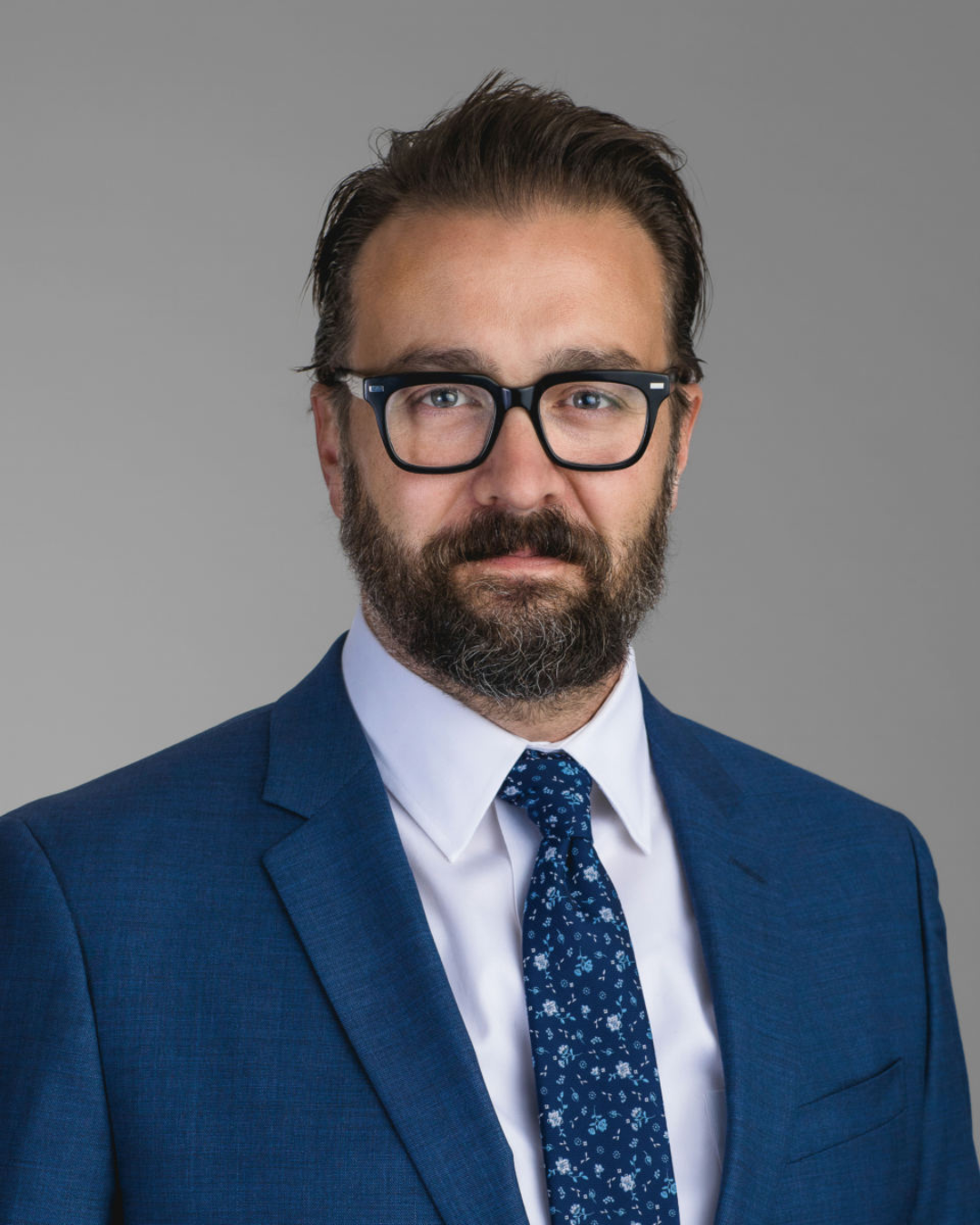 Man with short dark hair and dark beard wearing dark rimmed glasses, blue tie tie, and blue blazer looks into camera in front of light gray background