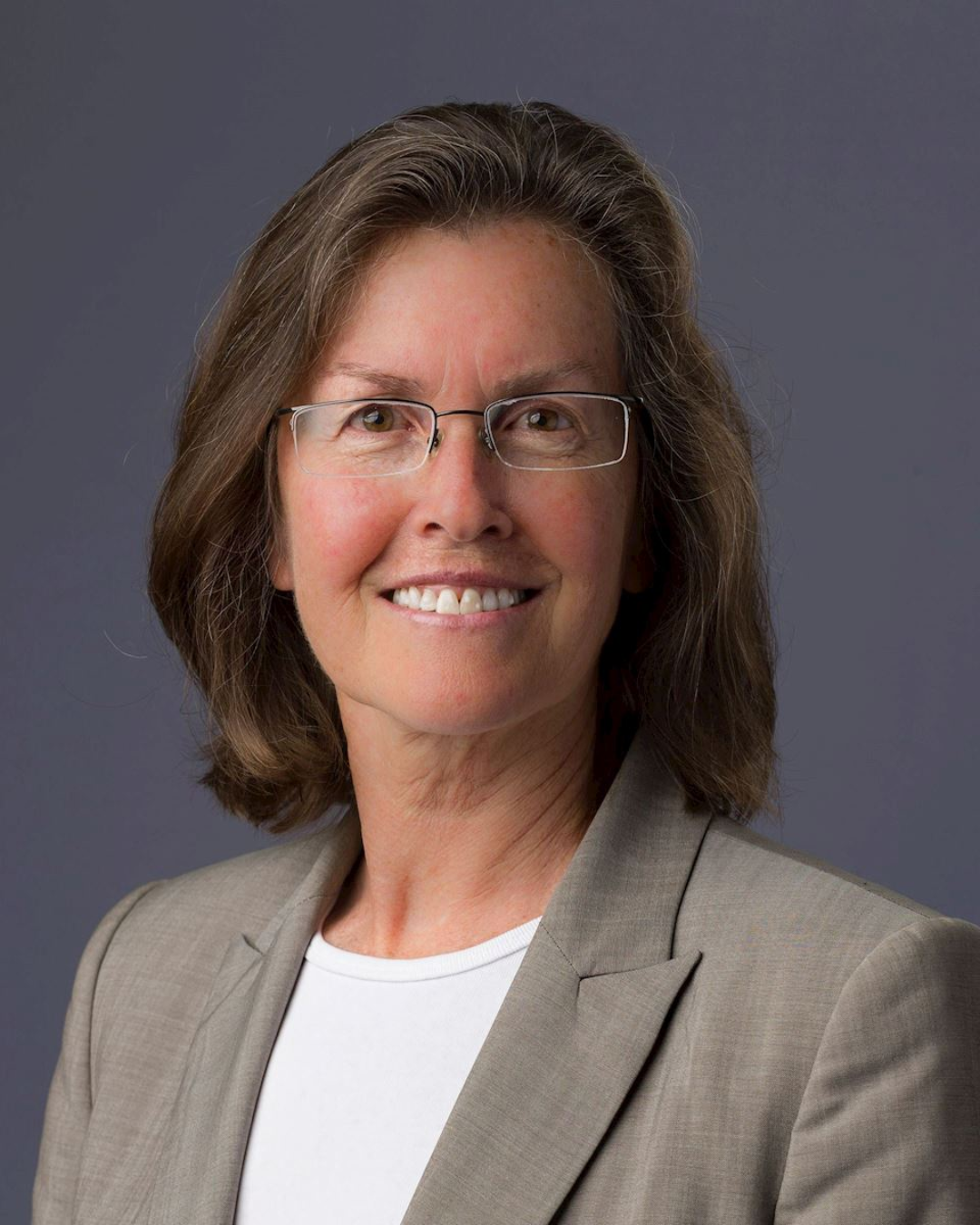 Woman with shoulder length brown hair and unrimmed glasses wearing tan blazer and white shirt smiles at camera in front of lavender background