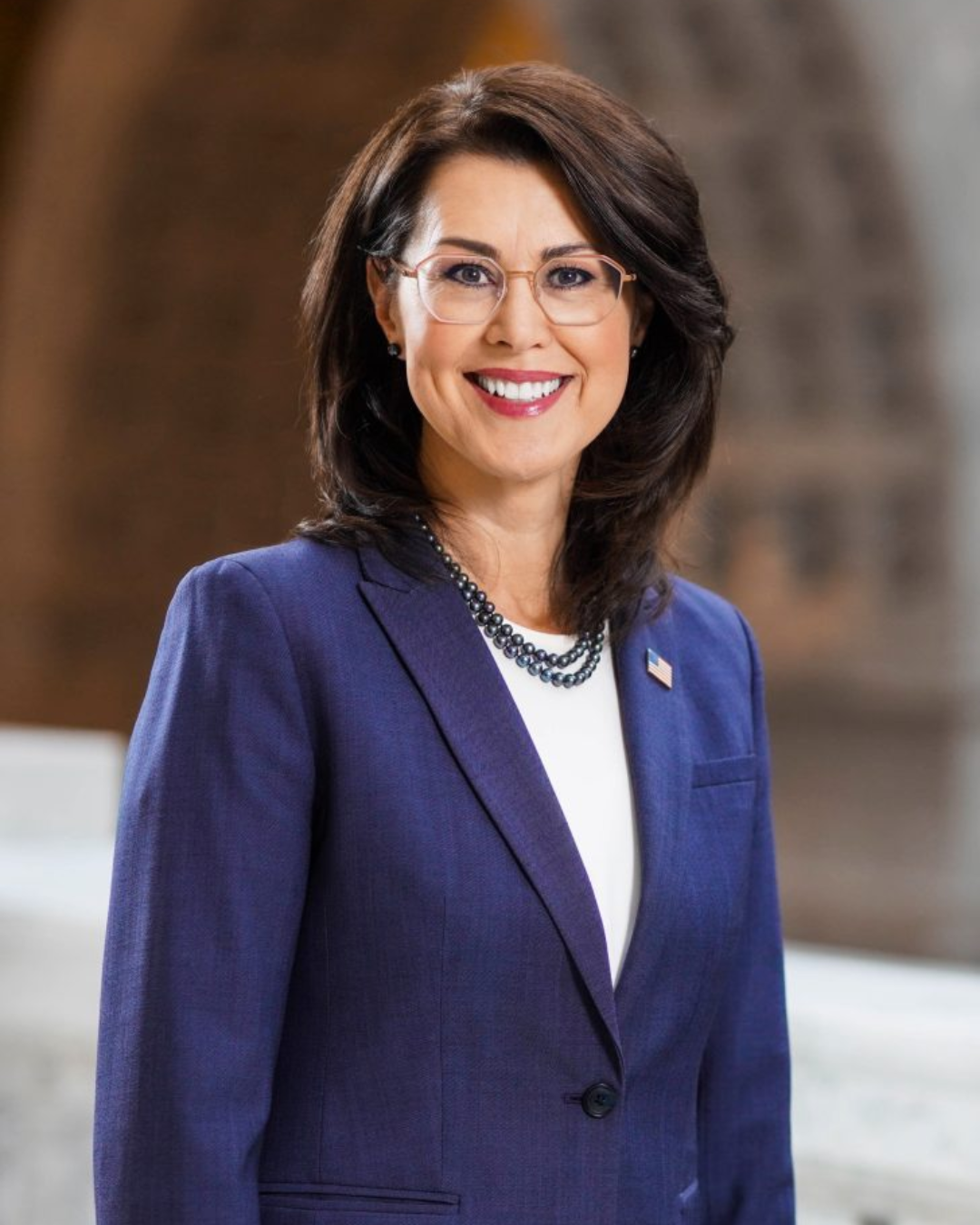 Woman with shoulder length brown hair and glasses smiles at camera wearing white top and navy blazer in the Utah Capitol building