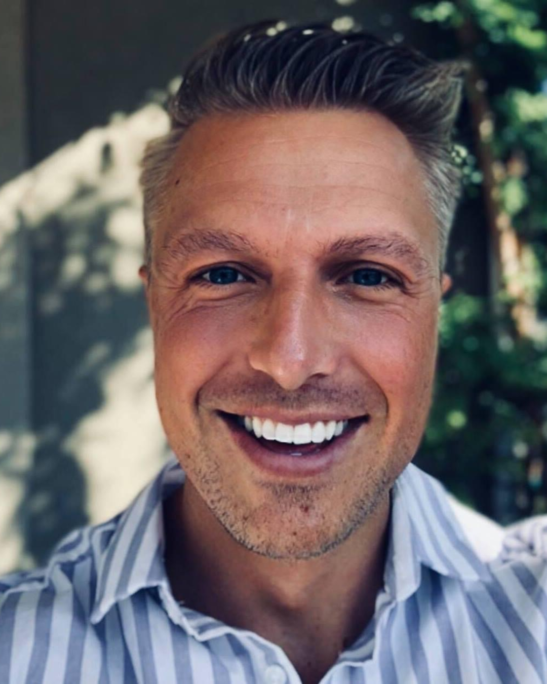Man with short hair smiles directly at camera wearing striped collared shirt in outdoor setting