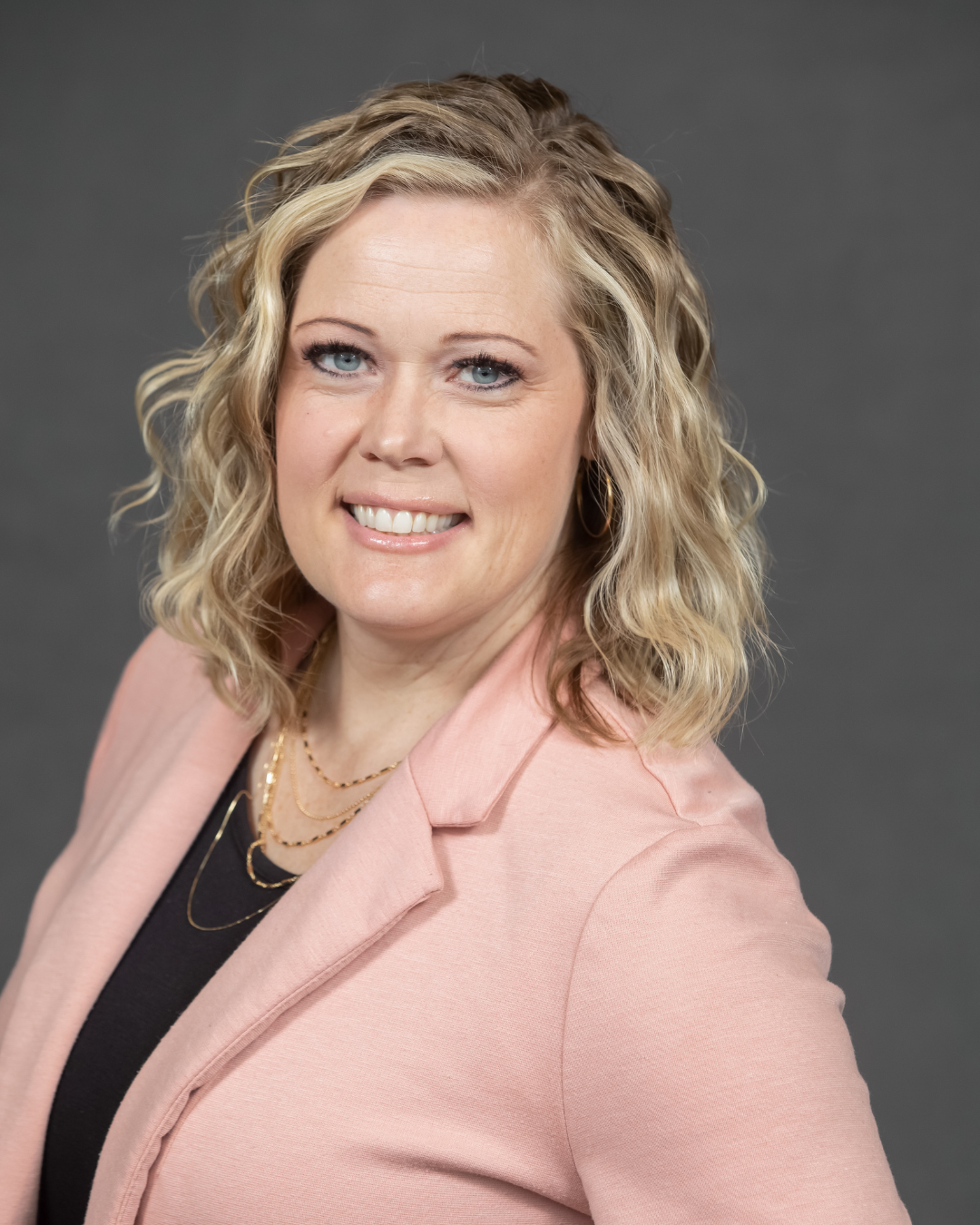 Woman with shoulder length wavy blonde hair smiles at camera wearing black top and light pink blazer in front of dark gray background