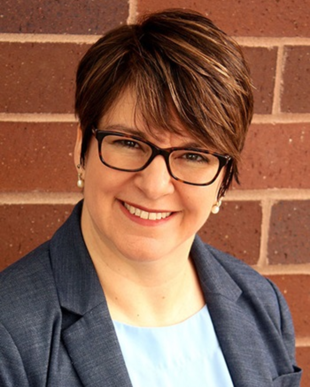 Woman with short brown hair and dark rimmed glasses smiles at camera wearing light colored shirt and dark gray blazer in front of red brick background