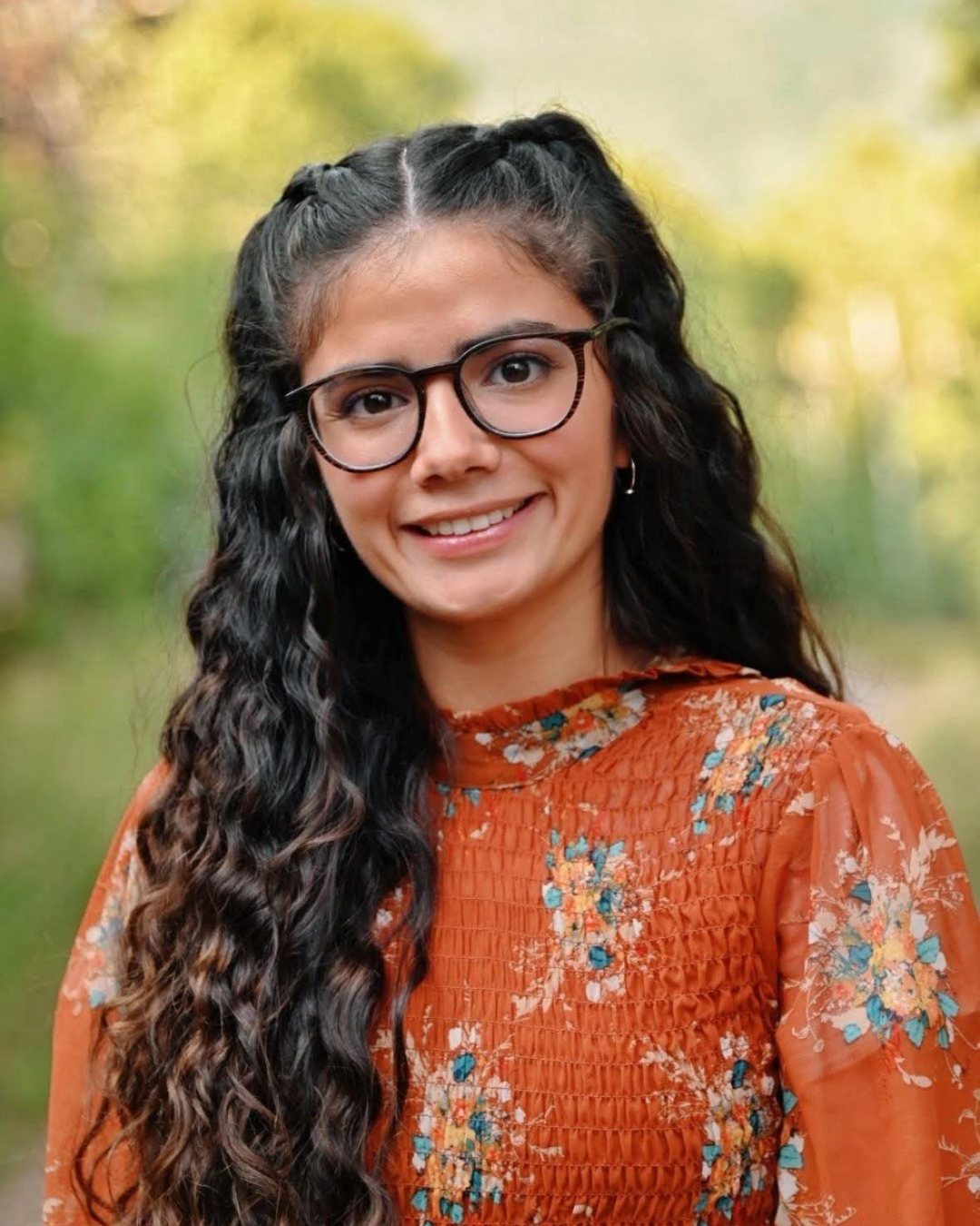 Young woman with long wavy dark hair smiles at camera wearing dark rimmed glasses and orange top with floral decorations in outdoor setting