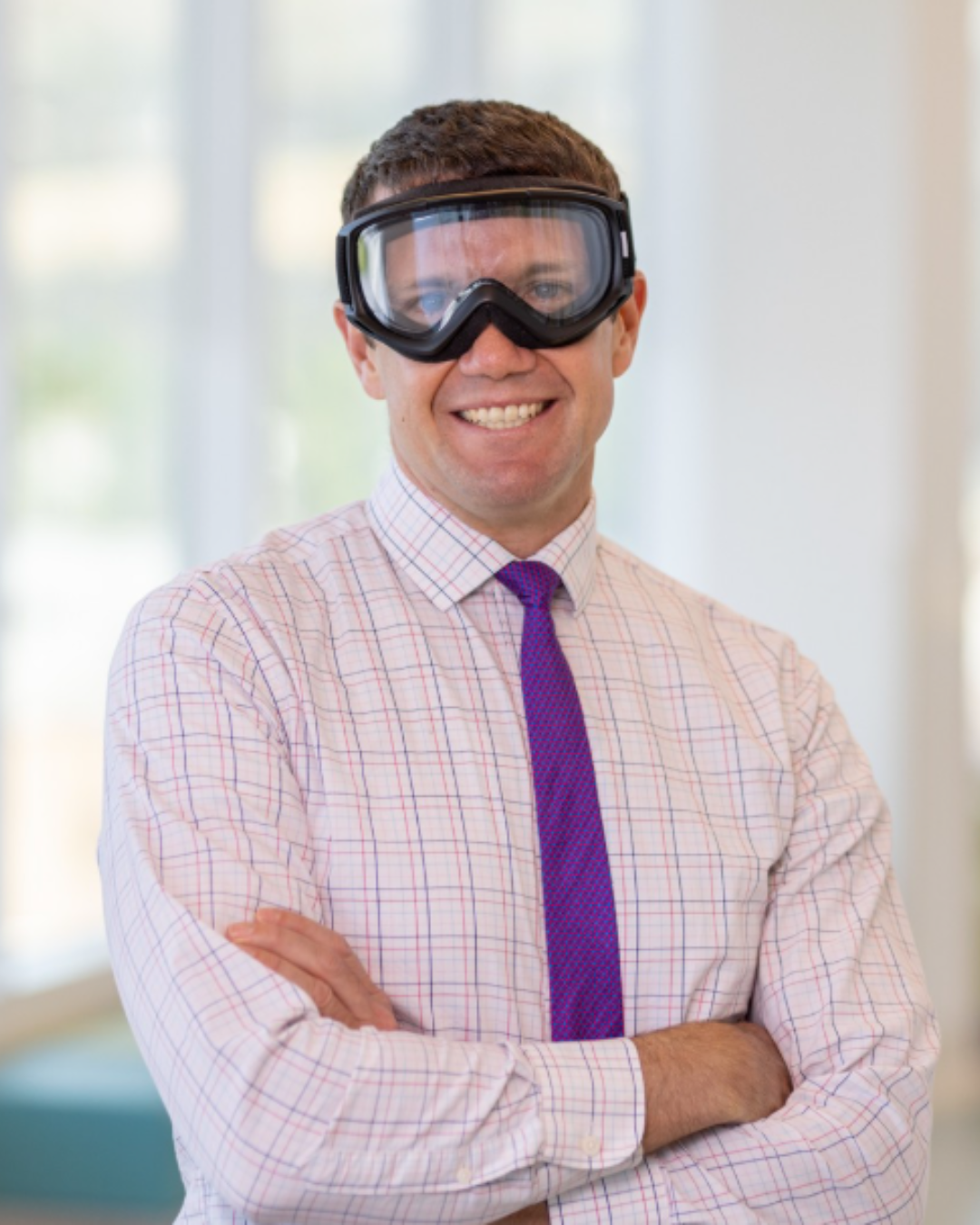 A man in a checkered shirt and purple tie smiles with arms crossed, wearing large safety goggles indoors.