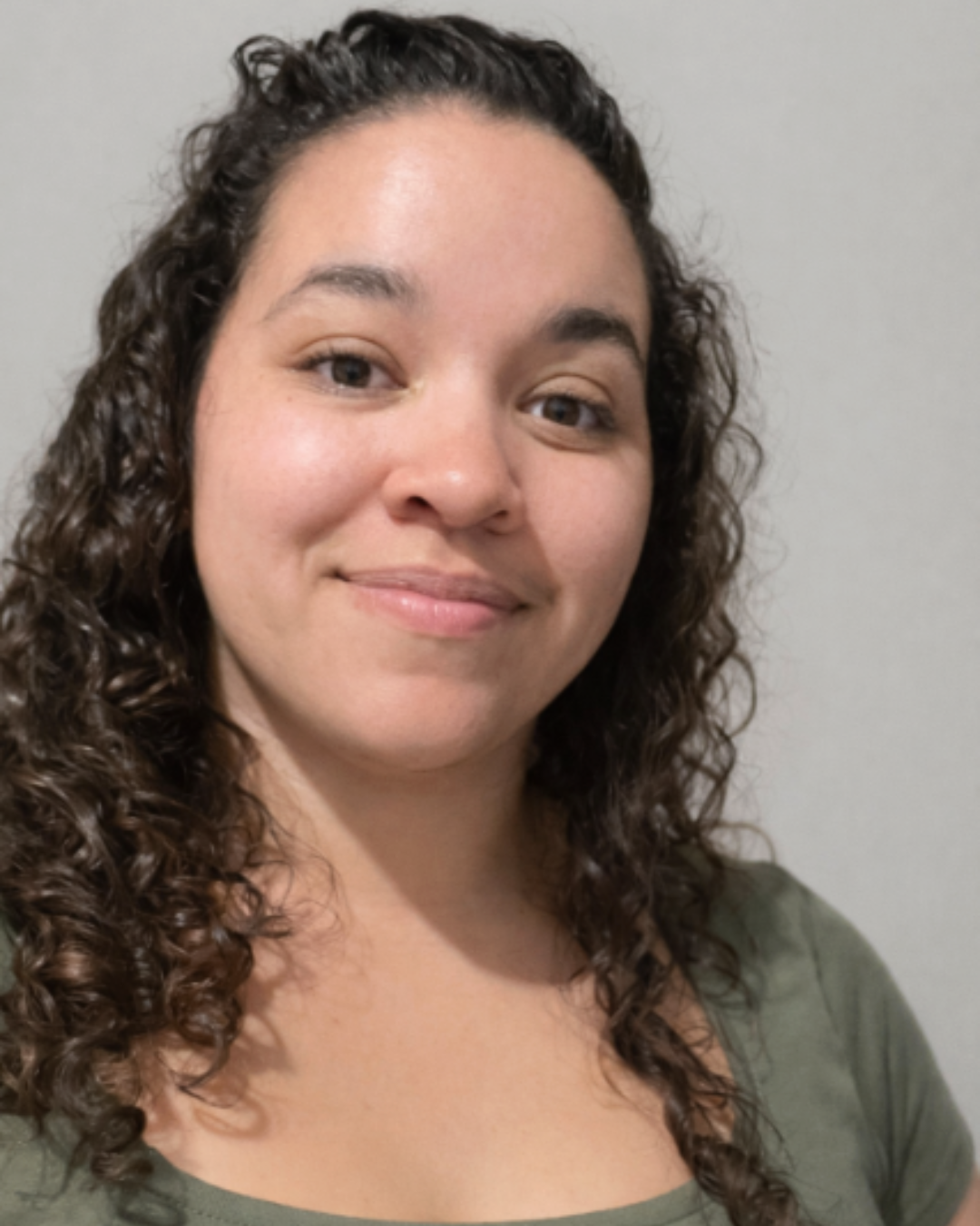 Woman with curly hair wearing a green top, smiling in front of a plain light background.