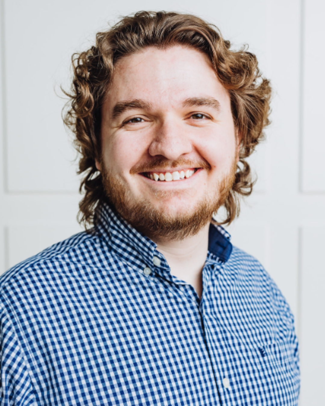 Smiling man with curly hair and a beard wearing a blue checkered shirt, standing against a bright background with a warm and friendly expression.