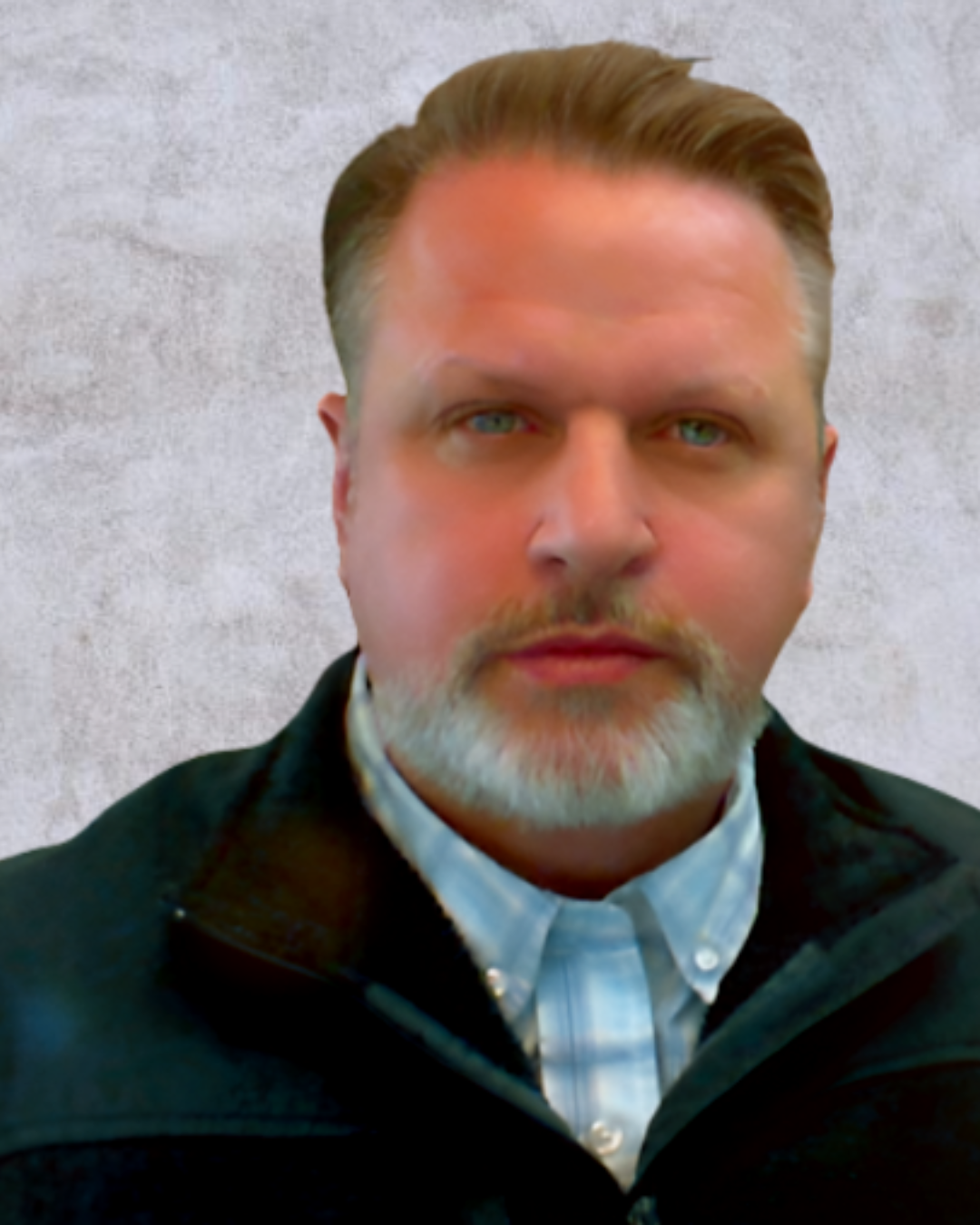 A middle-aged man with short brown hair and a trimmed beard looks directly at the camera. He wears a dark jacket and a plaid shirt against a plain background.