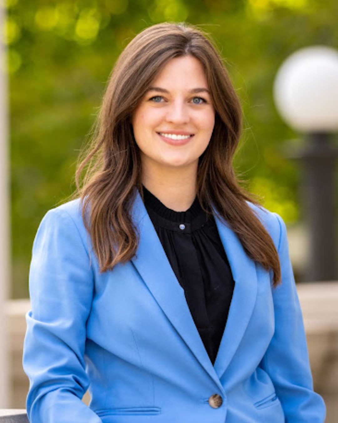 A young woman with long brown hair smiles at camera wearing black shirt and light blue blazer in outdoor setting