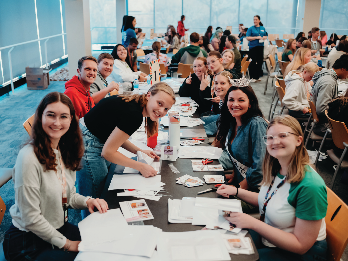 A group of people sit at tables assembling papers and smiling in a large, busy room.