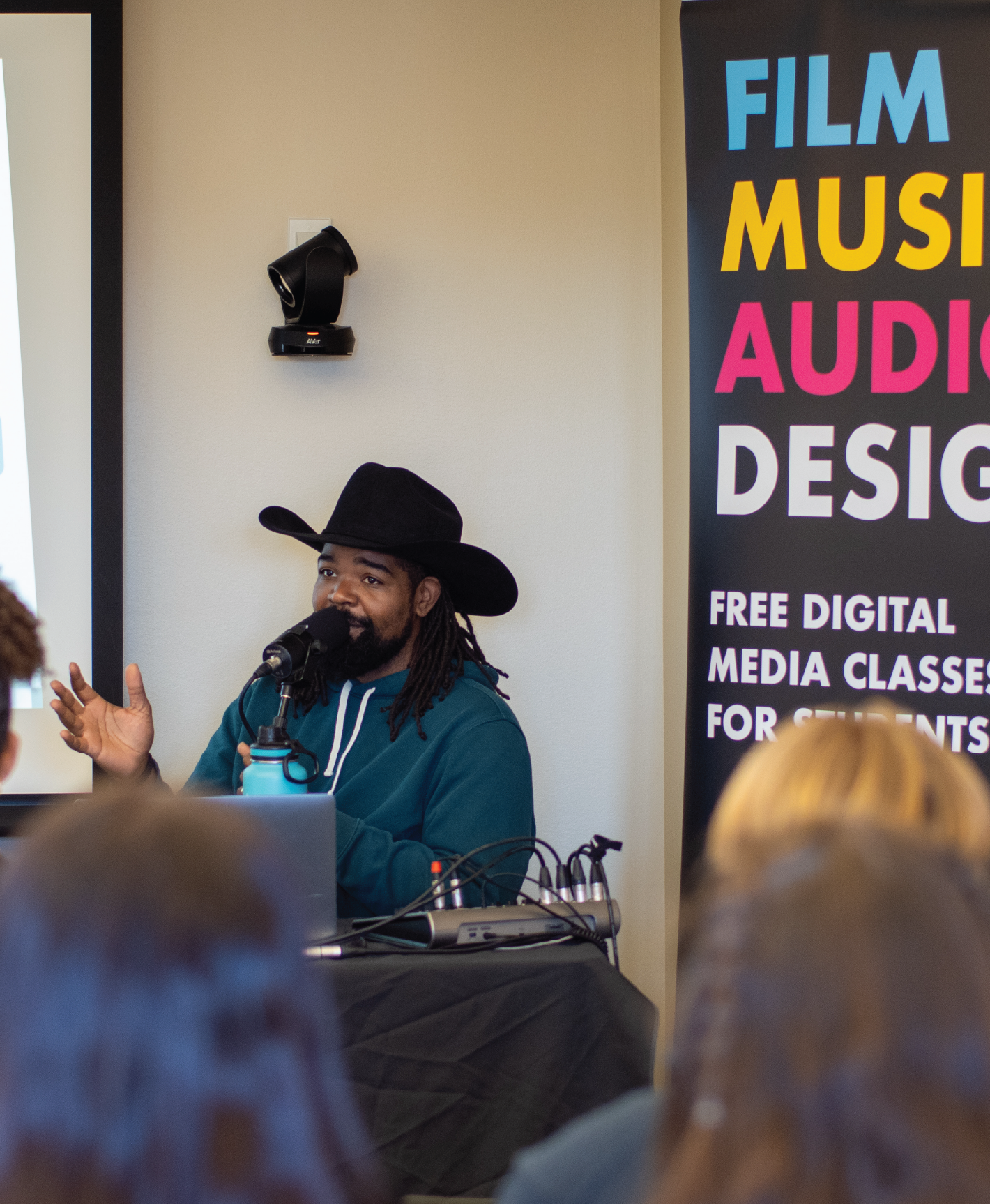 Man in a black cowboy hat speaks into a microphone at a media class event.