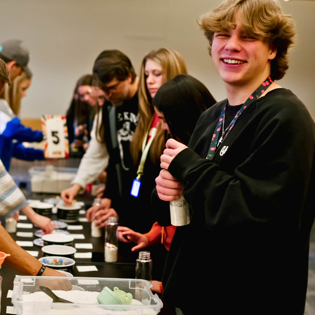 A group of young people make crafts at a table; one boy smiles at the camera.