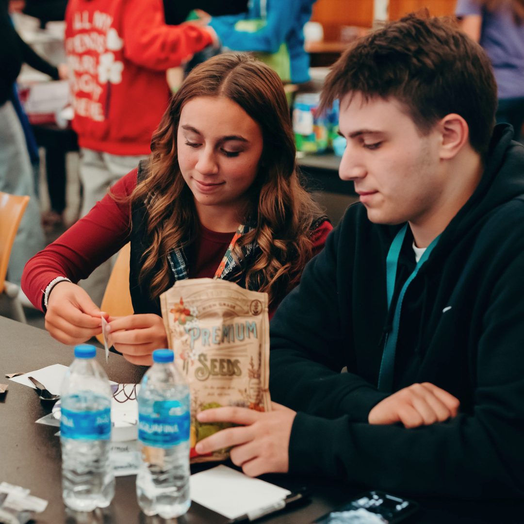 Two people sit at a table with water bottles and a bag labeled Premium Seeds in front of them.