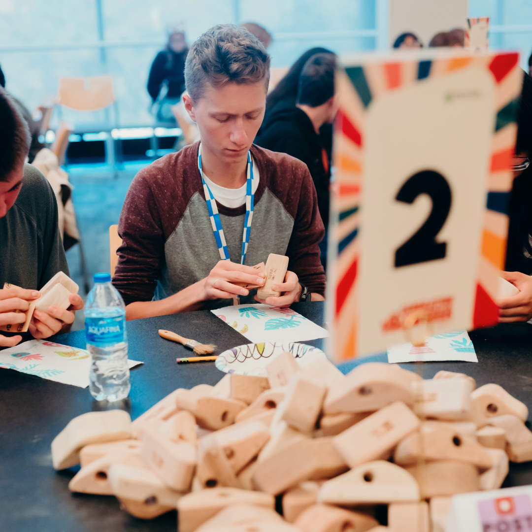 Person assembling wooden pieces at a table, with a 2 sign and water bottle visible.