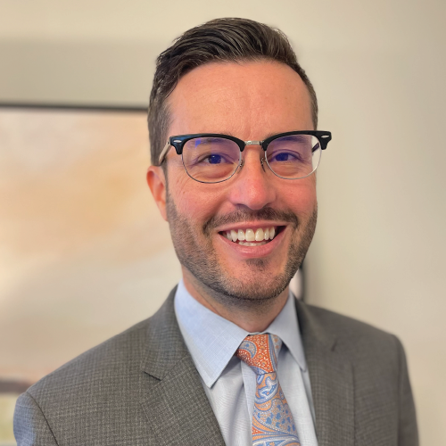 Man wearing glasses, a gray suit, blue shirt, and patterned tie, smiling at the camera.