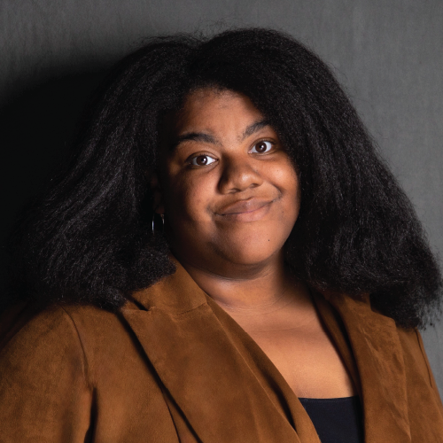 Person with long, dark hair in a brown jacket poses against a dark, neutral background.