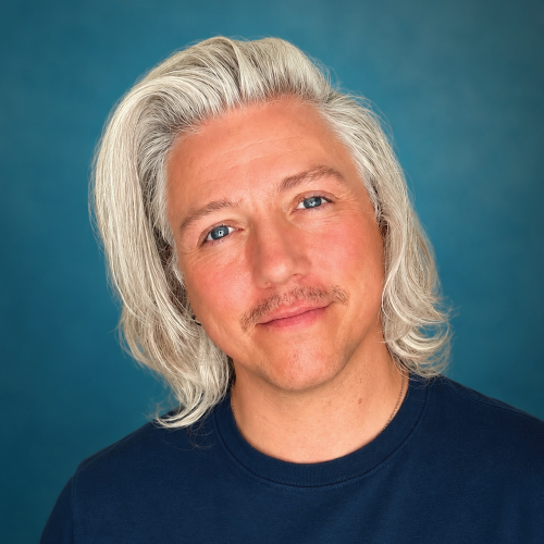 Smiling person with long, wavy gray hair and a mustache against a blue background.