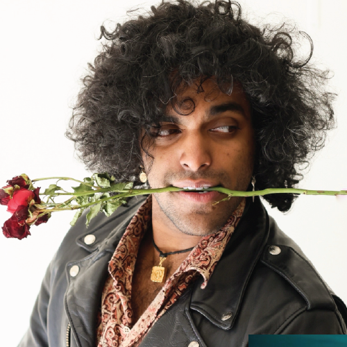 A person with curly hair wearing a leather jacket holds a red rose stem in their teeth.