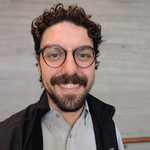 Man with curly dark hair, glasses, and a beard smiling at the camera against a gray wall.