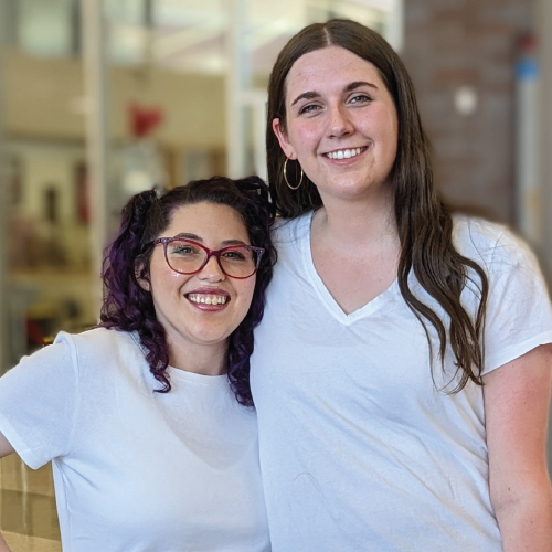Two women in white shirts stand indoors, smiling at the camera.