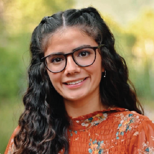 A young woman with long dark wavy hair and glasses wearing a floral top.